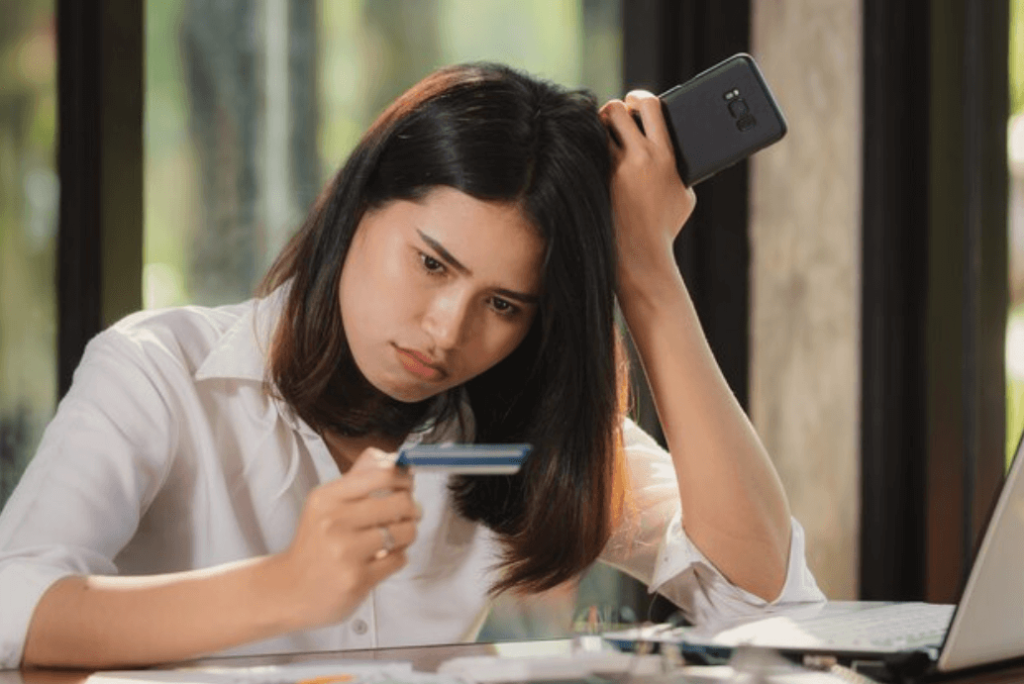 A concerned woman holding a credit card in one hand and a smartphone in the other, looking at the card with frustration while sitting at a desk with a laptop