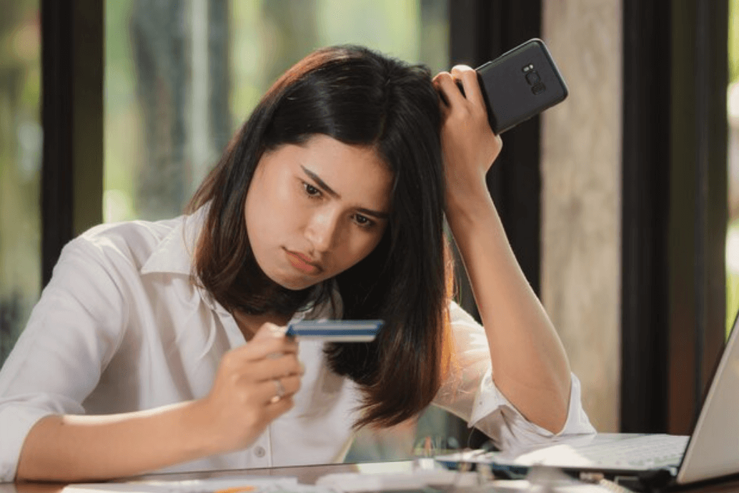A concerned woman holding a credit card in one hand and a smartphone in the other, looking at the card with frustration while sitting at a desk with a laptop